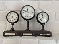 Front view of a metal clock with three round dials showing times for Los Angeles, New York, and London with black numbers and hands on white faces mounted on a rectangular base with timezone name plates.
