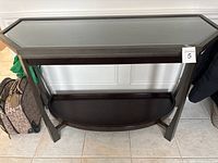 Front view of black wood console table with glass top and lower shelf, showing legs and shelf structure.