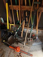 Garden tools assortment showing shovels, rakes, pitchfork, brooms, and electric hedge trimmer arranged against a garage wall