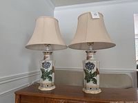 Pair of two cream-colored lamps with floral and butterfly designs on ceramic bases, placed on wooden surface.