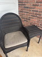 Outdoor black and brown wicker chair with beige cushion, placed next to a matching wicker table, located on a covered area with a brick wall background.
