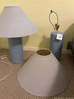 Two baby blue lamps shown, one with light gray shade attached and the other missing the shade and socket assembly; both with white cords attached on carpeted floor.
