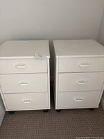 Front view of two white melamine night stands side by side with three drawers each and white handles, standing on caster wheels.