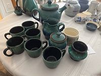 Top-down view of six green-blue glazed cups, teapot, creamer, and sugar bowl on a cloth-covered table showcasing the full tea set.