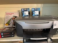 Front view of HP Photosmart 2610 printer with photo paper and ink cartridges on top and beside it