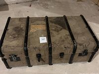 Front view of the vintage travel trunk placed on concrete floor showing fabric cover, black metal strips, locks, and wear marks.