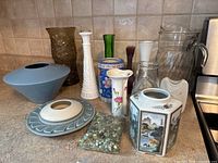 Top-down view of an assortment of 13 vintage vases and a bag of small pebbles on a kitchen counter.