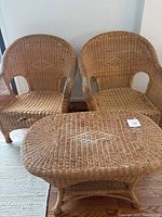 Two wicker armchairs and one wicker table grouped indoors on wood floor, set shows weathering and wear.