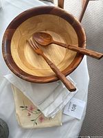 Wooden salad bowl with carved wooden salad fork and spoon laid on top, placed on folded table linens.