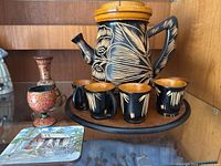 Full view of vintage carved wood water jug with lid and handle, four carved wood cups with black exterior and natural wood interior, small decorative carved wood pitcher or vase, coaster, and wooden tray, all displayed on a shelf.