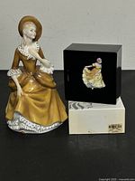 Photo of both Royal Doulton figurines placed side by side on black surface, showing the yellow dress of the larger "Sandra" figurine and the boxed miniature "Ninette" figurine.