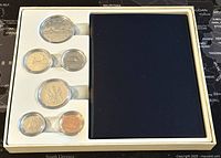 Top-down view of the coin set in the presentation case showing six coins embedded in clear holders and a navy blue cover on the right.