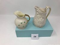 Two Belleek porcelain creamers displayed side by side, showing embossed patterns and handles