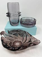Three amethyst-colored glassware items arranged together: an Art Deco vase, a decorative dish, and a humidor canister with a glass lid, all showing varied shapes and sculptural features consistent with early 20th century design.