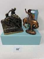 Two cast cowboy-themed items side by side with soda can for size reference. One is a cowboy on bucking horse figurine with raised arm; the other is a bookend with cowboy on horse with lowered head.