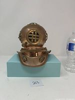 Front view of the copper-plated miniature diving helmet on a blue block, showing the central window with grill and flared neck base.