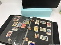 Open black binder with US postage stamps displayed on black pages inside plastic sleeves. Various denominations and designs visible.