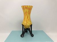 Front view of the tripod vase showing its yellow and orange spatter glass with black tripod base.