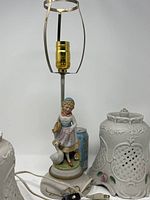 Porcelain lamp base showing figurine of a girl holding a basket with a white goose beside her, lamp socket without shade, and two white porcelain lamp shades with detailed floral and lattice designs nearby.