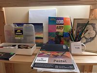Photo showing the arrangement of various art supplies on a shelf including paint brushes, colored pencils in a mug, a pastel pad, a sketch pad, a paint kit, and an art project book