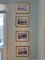 Four framed winter scene prints by Cornelius Krieghoff arranged vertically on a wall, showing the gold frames and white mats around detailed winter landscapes.