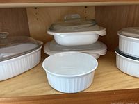 Six Corningware baking dishes of varying sizes arranged in a cabinet. White with ribbed vertical pattern. Four dishes have glass lids, one lid is missing. One dish has a plastic travel lid.
