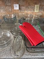 Overall view of glass bakeware including mixing bowls, pie plates, casserole dishes, gravy boat, and red oven mitts on kitchen countertop.