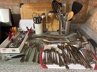 Full view of the kitchen counter showing bamboo cutting board, utensil holders with ladles, spatulas, kitchen scissors, various knives and forks in trays