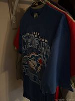 Blue 1992 American League Champions Toronto Blue Jays t-shirt hung on rack with other clothing items visible.