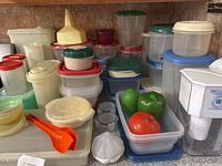 Image showing multiple plastic containers with lids in assorted colors, along with vegetable keeper containers with plastic vegetables inside and orange serving tongs.