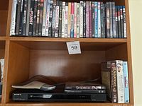 Photo showing Sony DVD player (DVP-NS78H) beneath a shelf filled with assorted DVDs, plus a few VHS tapes on the side.