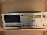 Front view of TEAC A-100 cassette deck showing VU meters, tape transport buttons, and controls.