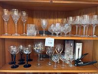 Full view of the shelves showing numerous crystal wine glasses, cognac snifters, tumblers, and wine accessories including a cork opener and boxed wine finer tools.