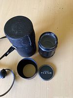 Photo of the Asahi Takumar 135mm f/3.5 lens, black metal body, with lens cap and leather carrying case on a light wood surface.