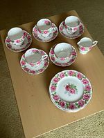 Top-down photo of 4 cup and saucer pairs, cream and sugar set, and a 7 inch plate all with pink rose floral design on white porcelain, displayed on wooden surface.