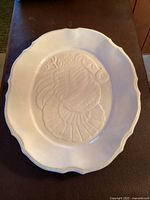 Top view of large white ceramic turkey platter with embossed turkey design in center and scalloped edged border.