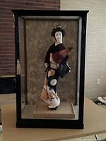 Front view of the Geisha doll in glass display case showing detailed face, kimono, and red fan.