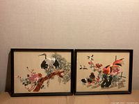 Pair of vintage silk embroidered artworks mounted in rectangular dark wooden frames resting against beige wall