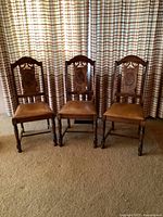 Three vintage oak wood chairs with carved backs and vinyl seat covering, positioned side by side in front of patterned curtains.