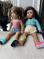 Two vintage American Girl dolls seated side by side on a surface, showing their full bodies with clothing and footwear.