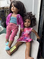 Two American Girl dolls, one standing and one sitting, shown with detailed clothing and hair styles, indicating condition and diversity.