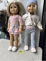 Two vintage American Girl dolls standing upright, one in a pink dress with a silver belt and pink shoes, the other in a white jacket, pink shirt, grey leggings, white shoes, and a blue beanie.
