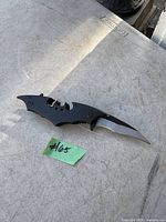 Photo of a black folding knife shaped like the Batman logo with a sharp, curved blade extending from the end of one wing, placed on a metal table with a green lot number tag.