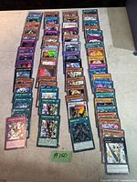 Top down view of about 70 Yu-Gi-Oh! trading cards laid out in six rows showing various monsters, spells, and traps from Konami's Yu-Gi-Oh! series.