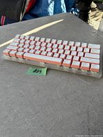 Photo of compact mechanical keyboard with white keycaps and orange accents on a gray surface, showing side angle and overall aesthetic.