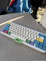 Mechanical keyboard with a white case and multi-colored keycaps, showing various colors such as blue, green, purple, yellow, and red arranged on a compact layout. The keyboard is photographed on a gray surface outdoors with a label #25 attached to its front.