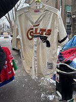 Front view of the white Mitchell & Ness baseball jersey with 'Colts' and revolver graphic, sleeve stripe, and button-up front.