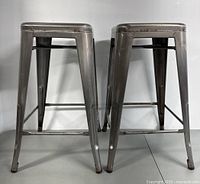 Front view of two metal bar stools side by side showing tapered legs and footrests.