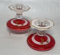 Pair of vintage Indiana Glass candlesticks with red bases and clear candle holders showing gold leaf vine decorations and gold trimmed edges.
