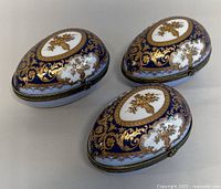 Three ceramic egg-shaped trinket boxes, cobalt blue and gold design, metal clasps visible, top view showing gold floral motif on white oval background.